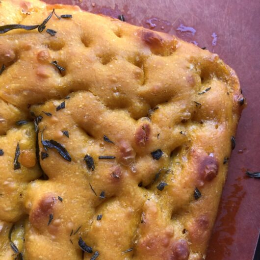 a closeup of a pumpkin colored corner of Delicata squash focaccia bread dimpled with bits of fresh rosemary baked on top