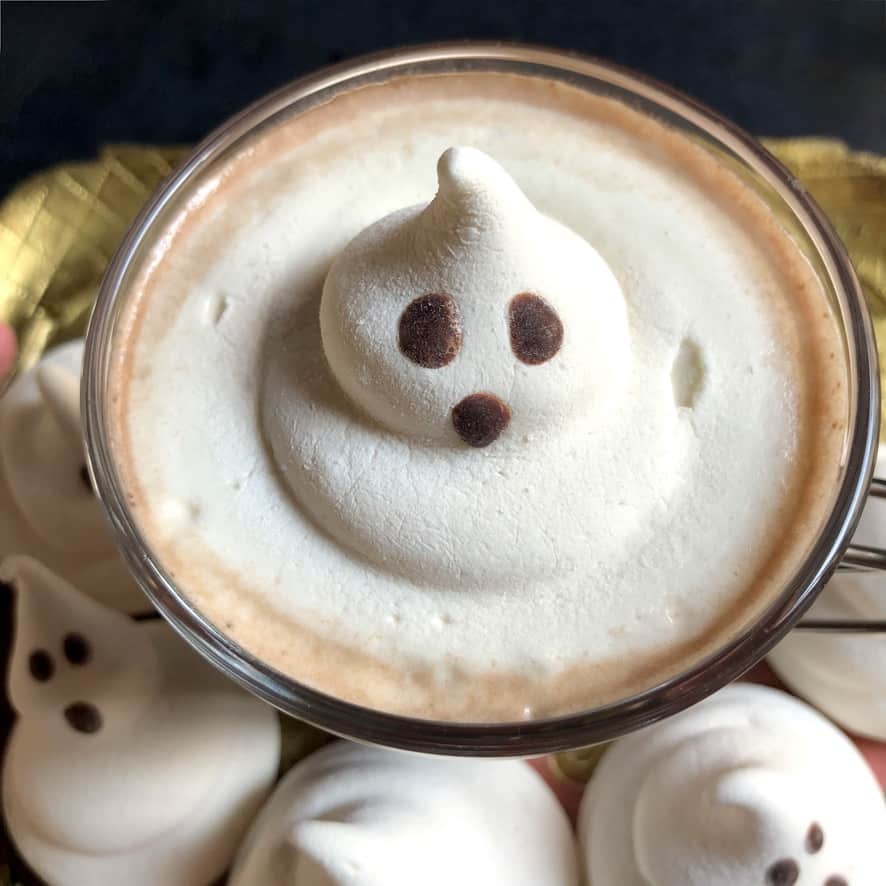 a ghost marshmallow slowly melting in a homemade hot cocoa surrounded by ghost marshmallows at the base of the barista glass mug