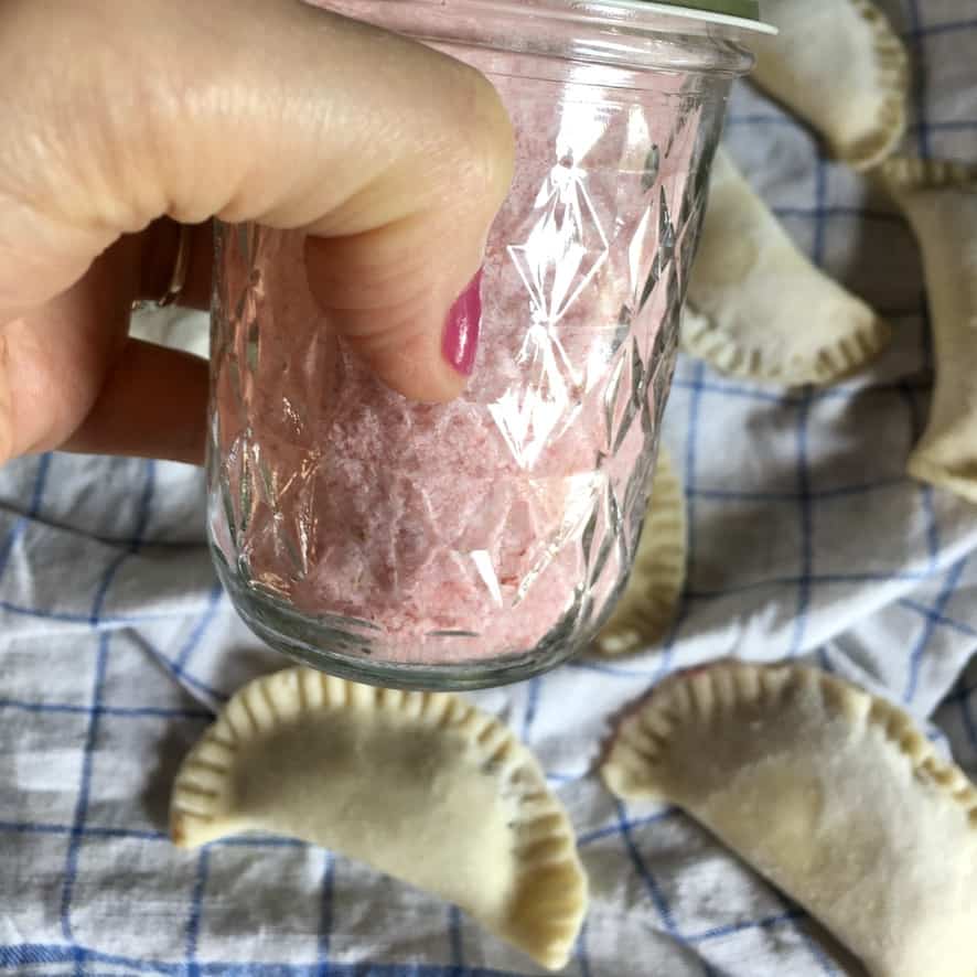 my hand holding a jar of strawberry sugar above a linen towel with uncooked fried pies.