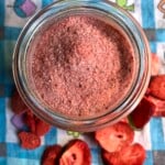 Deep dark red strawberry sugar in a jar with freeze dried strawberries scattered around it on a turquoise blue checked tea towel.