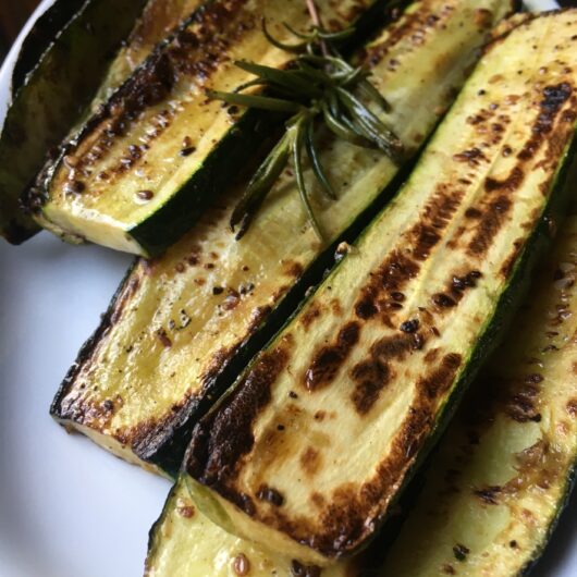 closeup pan-seared caramelized and golden brown zucchini slices on a white platter and a sprig of crispy rosemary on top