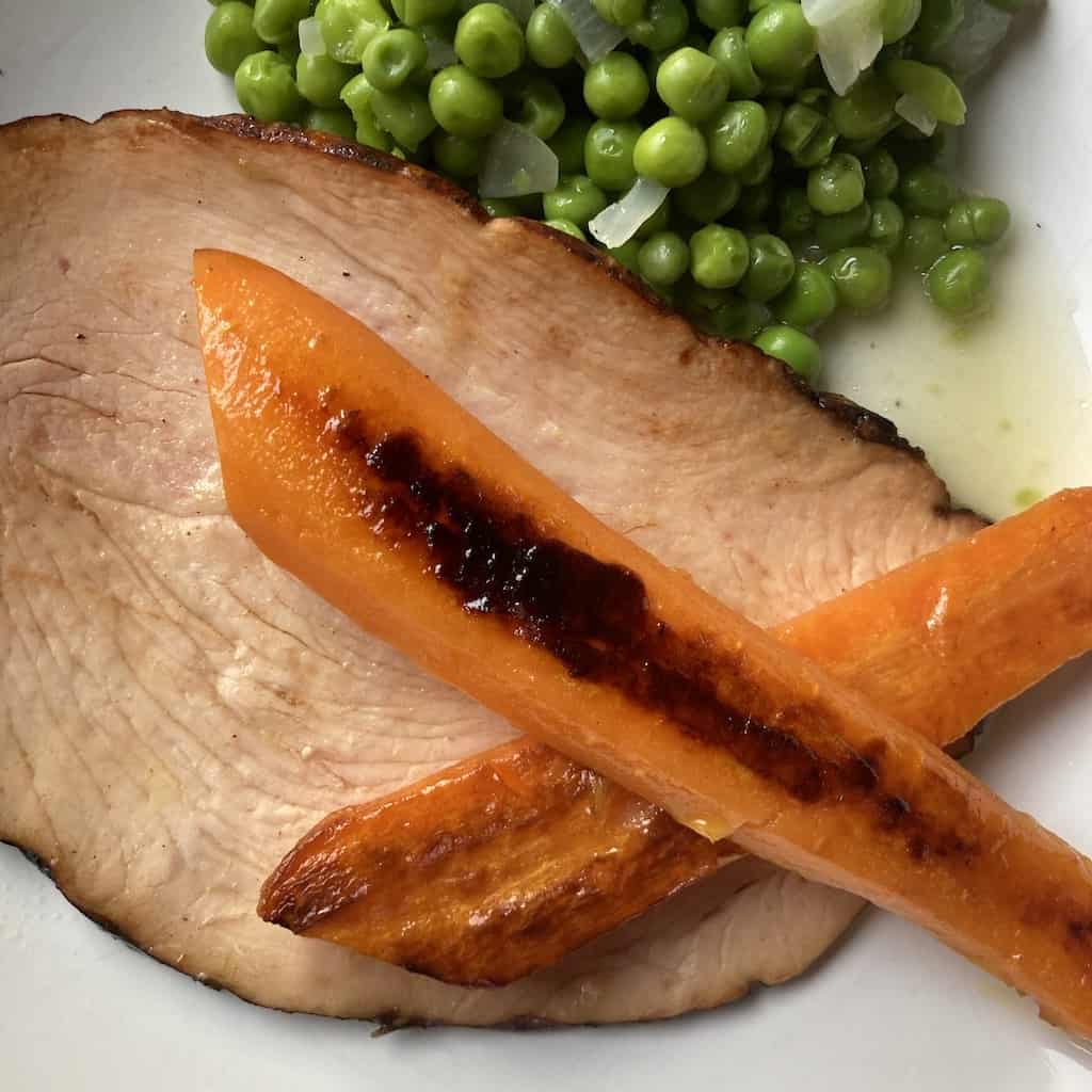 2 pieces of pan seared carrots criss-crossed over each other lying on top of a piece of roast turkey breast with a side of peas on a plate