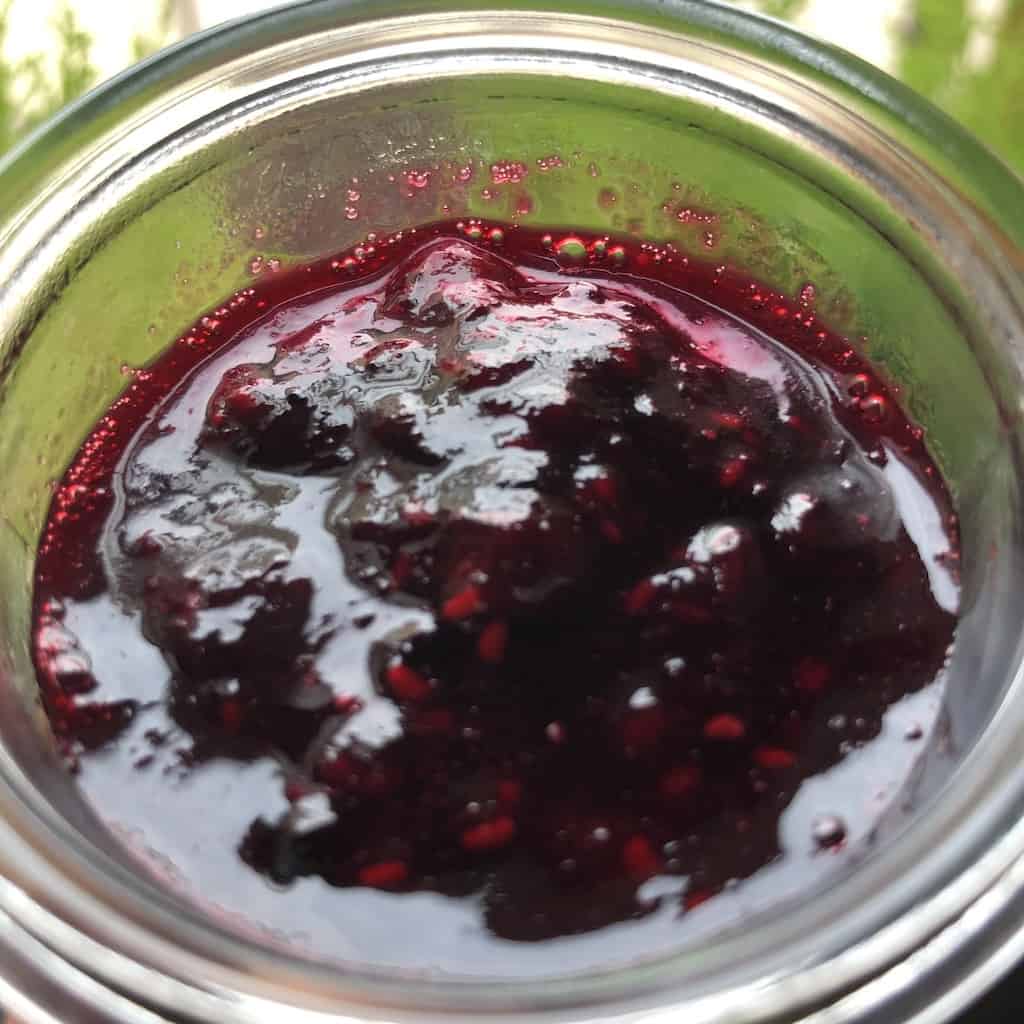 homemade berry fruit sauce in a spoon above the glass jar filled with bright red and blue sauce