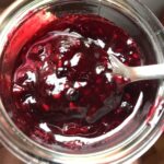 homemade berry fruit sauce in a spoon above the glass jar filled with bright red and blue sauce