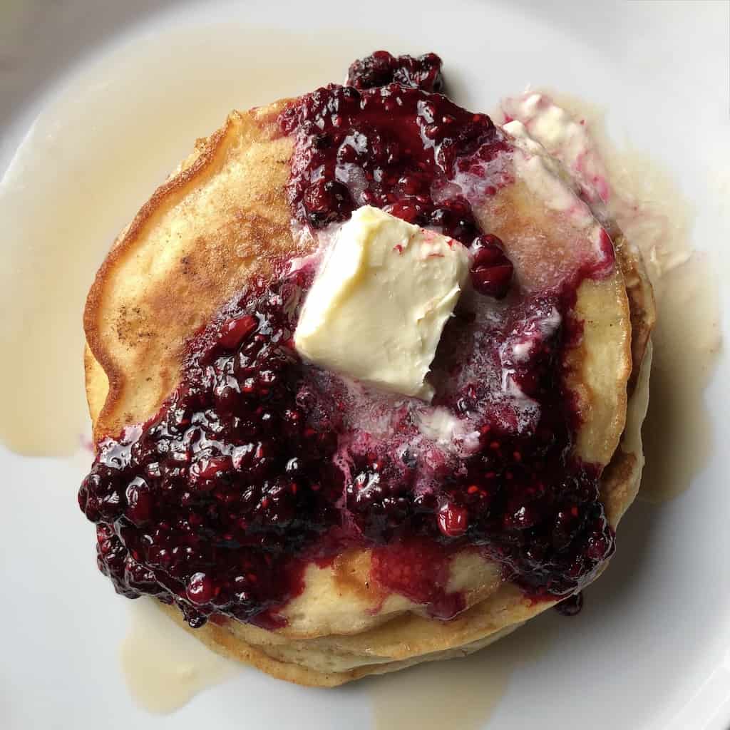 stack of beautifully fluffy golden pancakes with a fat pat of butter melting in the middle of a homemade berry sauce dripping on 3 sides and maple syrup oozing off the pile of pancakes creating a pool around the pancakes