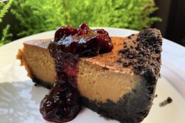 slice of an amber colored white chocolate caramel cheesecake with Oreo cookie crust drizzled with a beautiful homemade red mixed berry sauce with a view of bright fluorescent green mini cypress trees in the immediate background