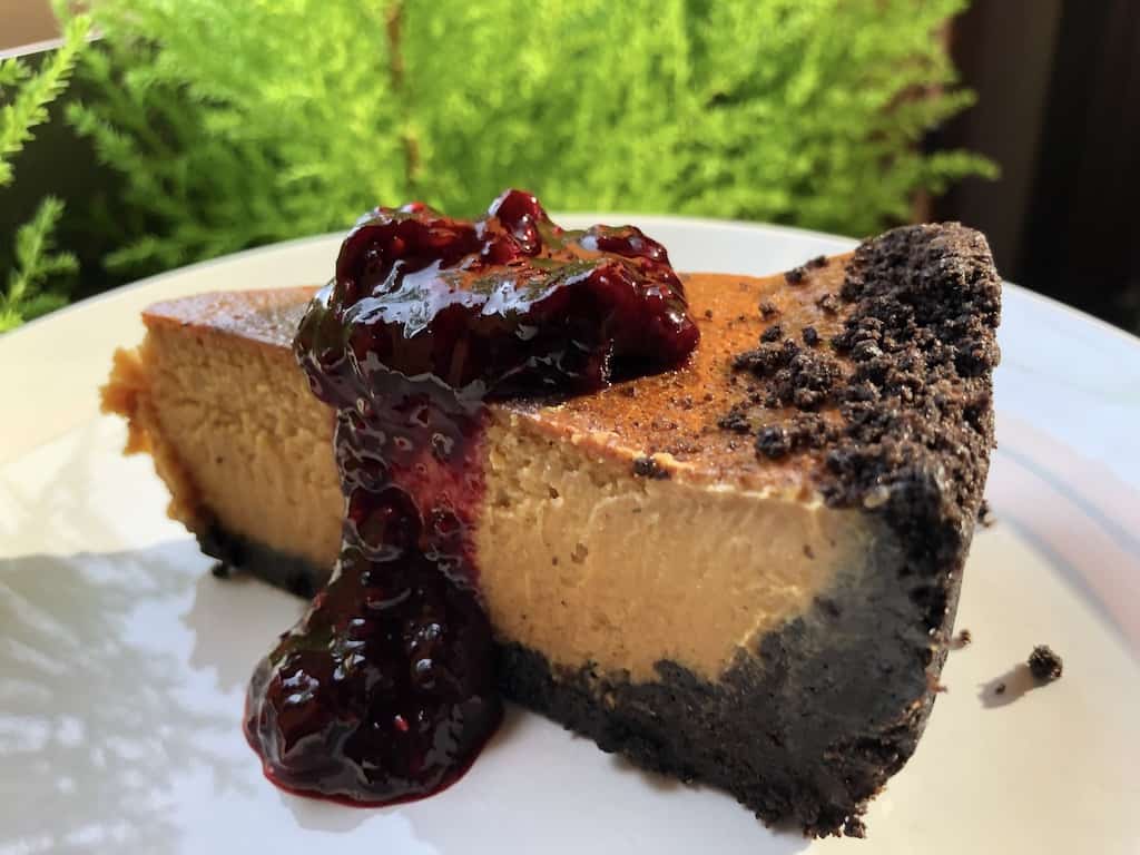 slice of an amber colored white chocolate caramel cheesecake with Oreo cookie crust drizzled with a beautiful homemade red mixed berry sauce with a view of bright fluorescent green mini cypress trees in the immediate background