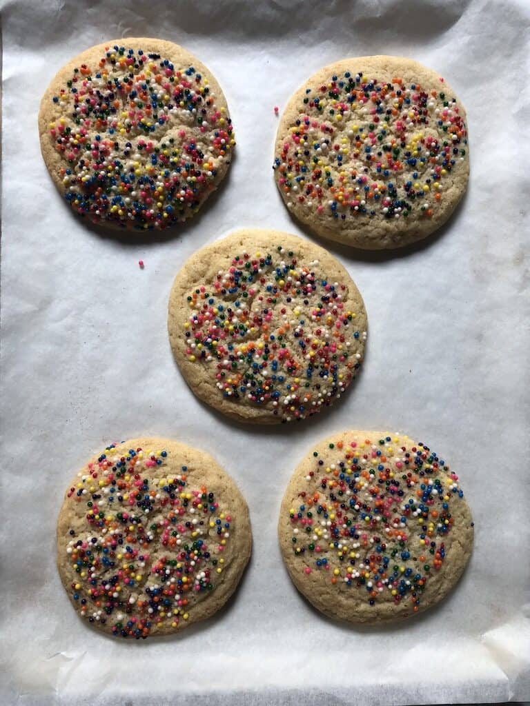a sheet pan lined with white parchment paper with freshly baked sugar cookies with nonpareil sprinkles