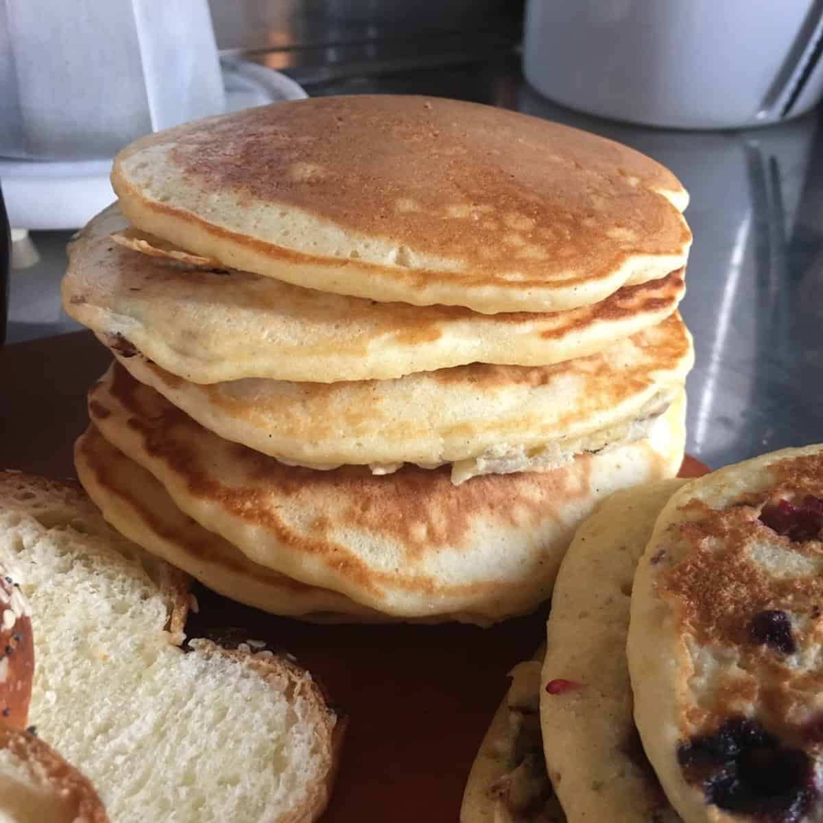 A tall stack of golden brown buttermilk pancakes from scratch next to blueberry pancakes and sliced homemade bread.
