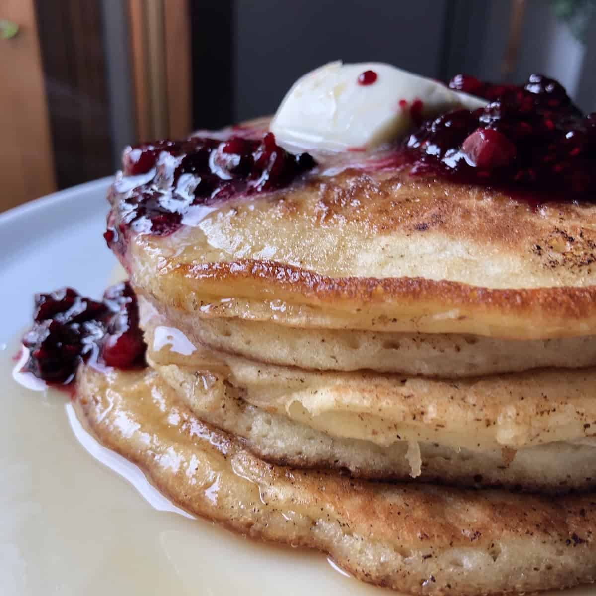 Closeup of a stack of buttermilk pancakes with crispy buttery edges topped with homemade blueberry sauce, maple syrup, and a knob of melting butter.