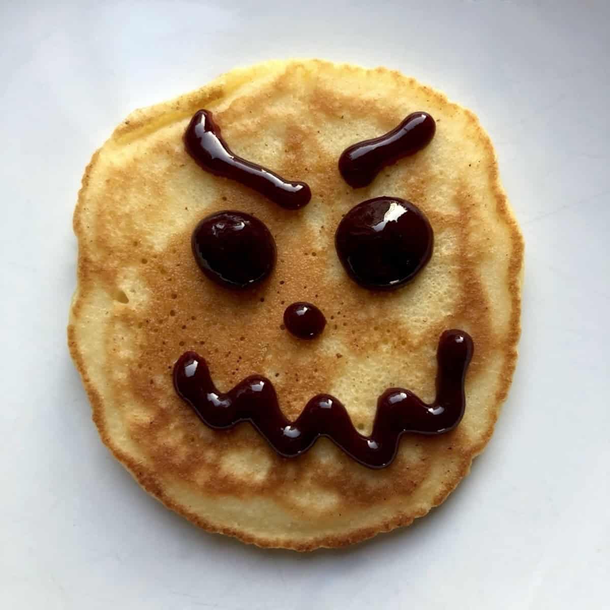 A golden buttermilk pancake with a jack-o-lantern face drawn in blueberry sauce for a fun homemade pancakes treat.