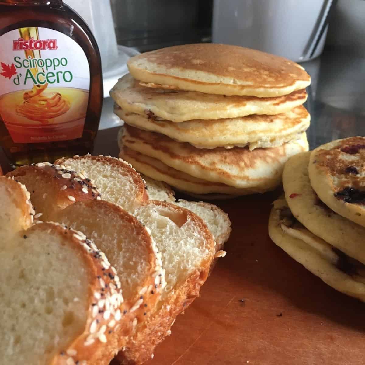 Stacks of hot fluffy pancakes with homemade slices of potato bread, a bottle of pure maple syrup, and pancake variations like blueberry buttermilk pancakes as well.