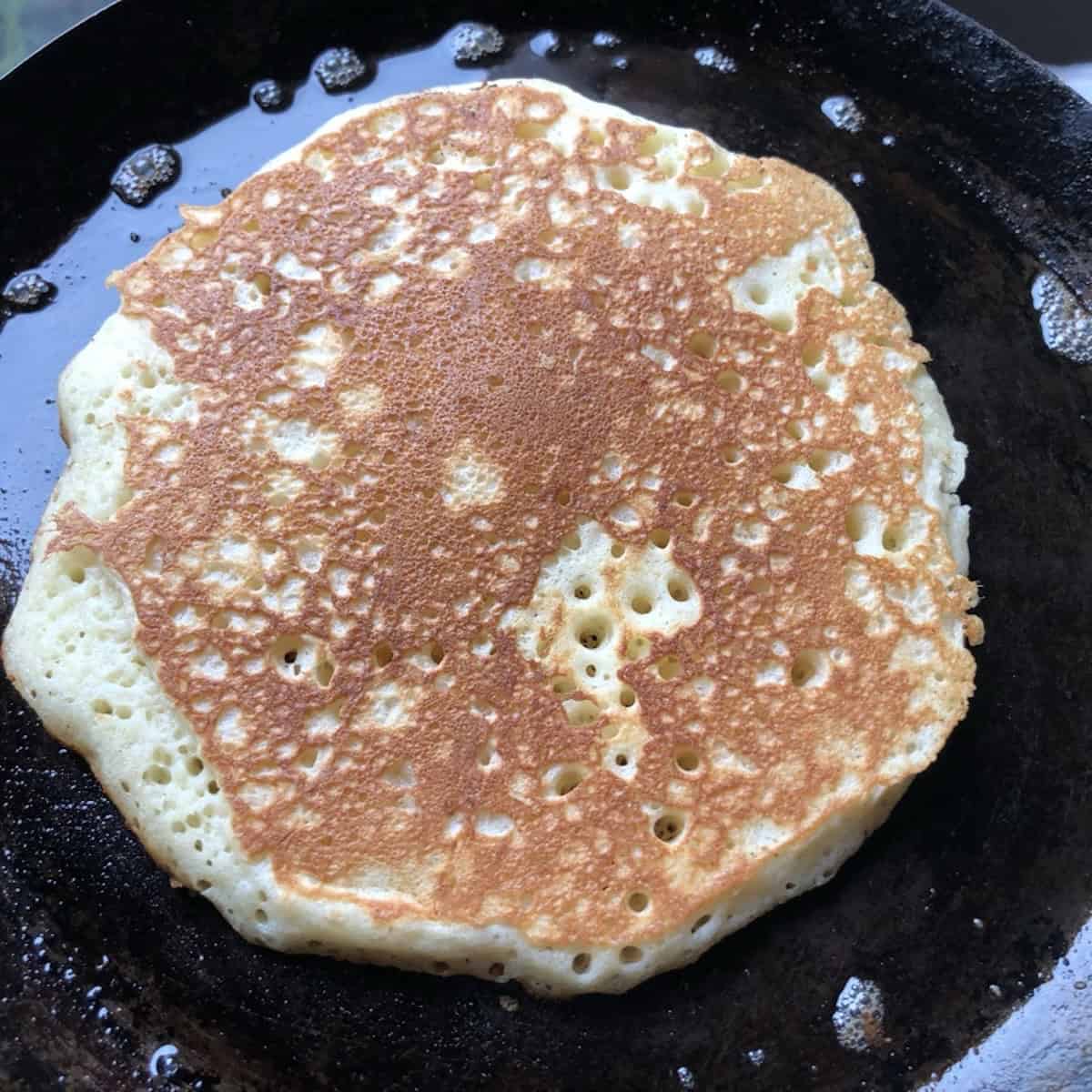 A golden brown pancake with a lacy browning pattern cooking in a buttery cast iron skillet.