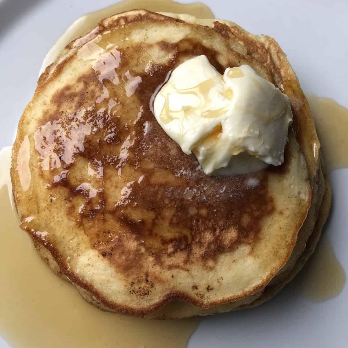 Top down view of a stack of buttermilk pancakes with melting butter and maple syrup on a white plate.