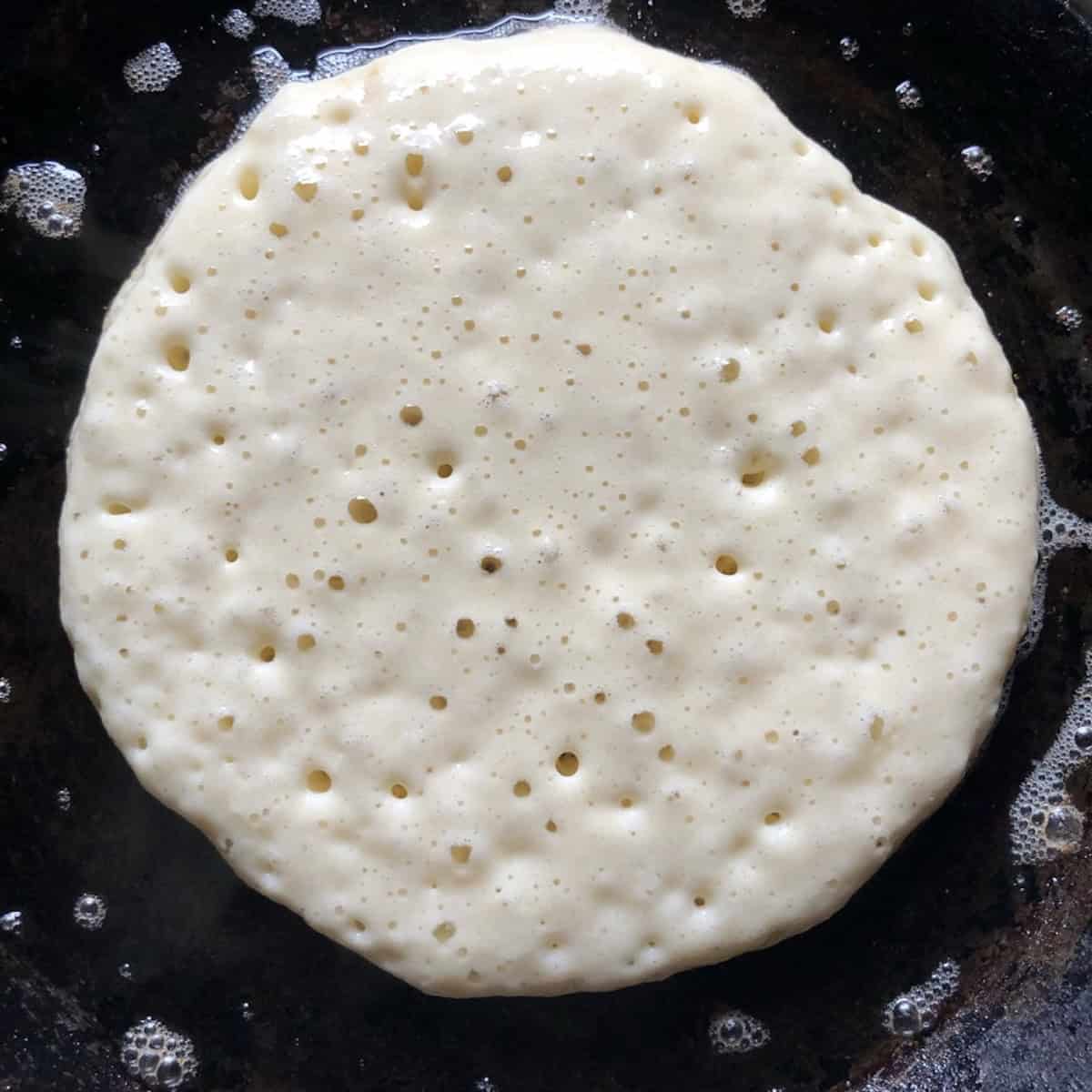 A buttermilk pancake cooking in a skillet with bubbles formed across the entire surface indicating it is ready to flip.