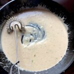 the perfect Alfredo Sauce in a cast iron skillet with a coil whisk showing the silky texture of the sauce