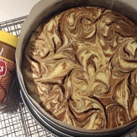 Delicious looking swirled Biscoff Cheesecake with a jar of Biscoff Crunchy Biscuit Spread lying next to the cheesecake still in the springform pan resting on a stainless steel cooling rack