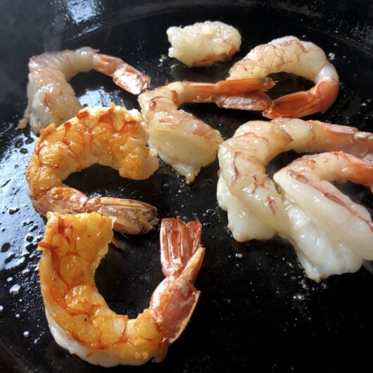 2 cooked shrimp that have been flipped over to reveal its natural orange-red color with crispy bits in contrast to the remaining shrimp that haven't yet been flipped over and are pale pink and white in comparison