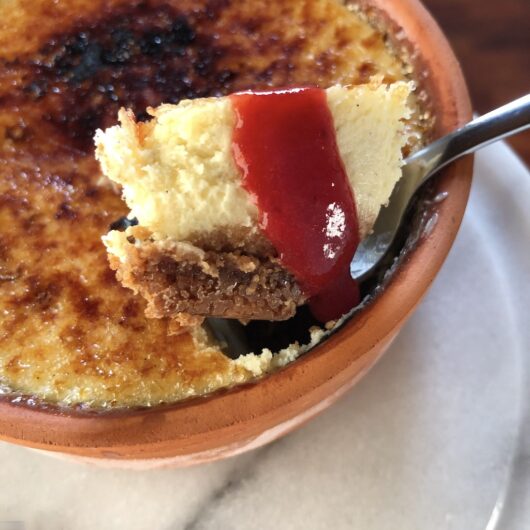 a bite of cheesecake in a small spoon and drizzled with strawberry sauce and a biscoff crust with the rest of the cheesecake in an individual terra cotta ramekin typically used for creme cramel