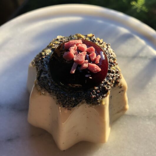white chocolate vanilla bean panna cotta in the shape of a Pandora (Italian star mold) with the poppy seeds and lemon zest concentrated at the top of the single panna cotta which has a deep berry red coulis sauce on top and chopped pink white chocolate