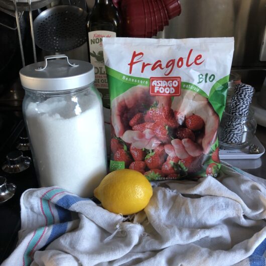 raw ingredients for the strawberry sauce on top of the counter including a bag of frozen strawberries, jar of sugar, and a fresh lemon