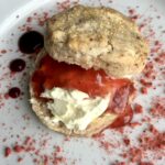 strawberry scone with homemade clotted cream and a scoop of strawberry sauce topped with the scone top and the plate dusted with pureed freeze-dried strawberries and three dots of strawberry sauce