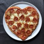 a baked heart-shaped pizza on a white ceramic round pizza platter on a grey linen tablecloth