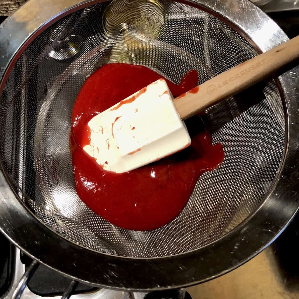 a white rubber le creuset spatula pressing down on the hot, pureed strawberry sauce that's in a sieve strainer in order to remove seeds and any large pieces