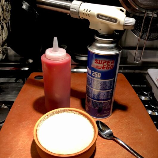 strawberry coulis placed in a white plastic squeeze bottle for plating next to an Iwatani Brand torch attached to a bottle of butane with a just-sugared white chocolate creme brùlée cheesecake in a terra cotta colored creme caramel dish