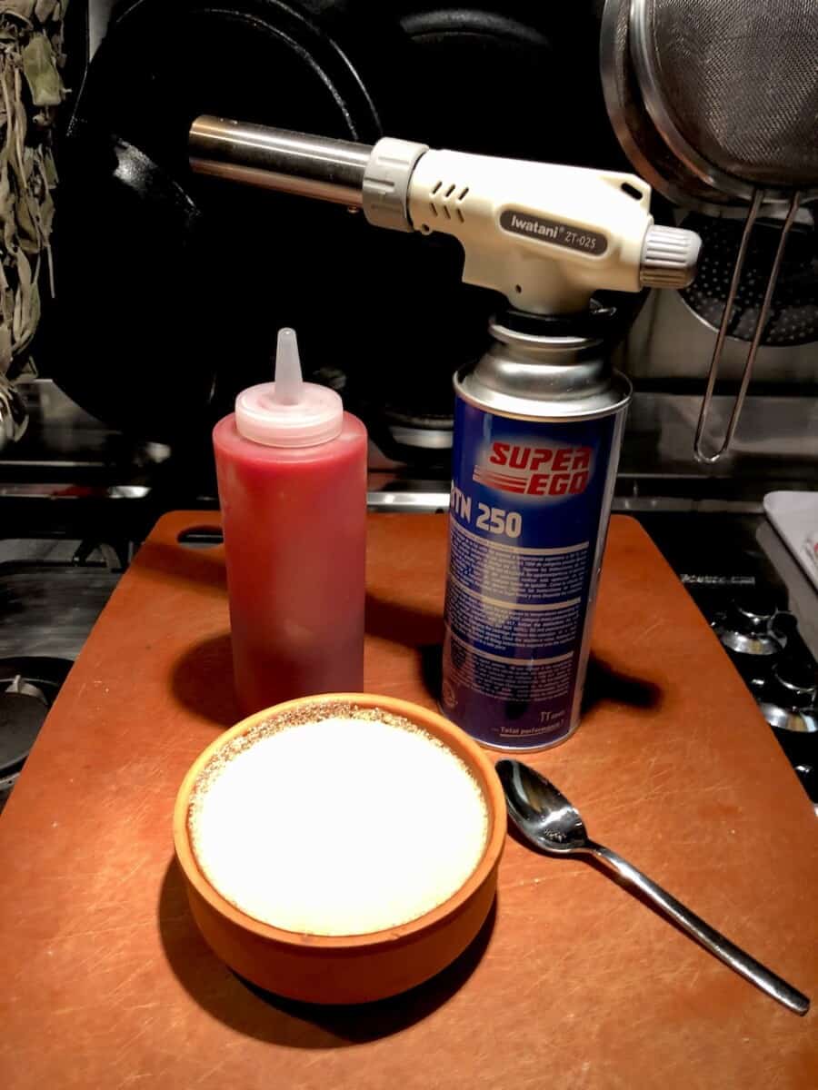 strawberry coulis placed in a white plastic squeeze bottle for plating next to an Iwatani Brand torch attached to a bottle of butane with a just-sugared white chocolate creme brùlée cheesecake in a terra cotta colored creme caramel dish