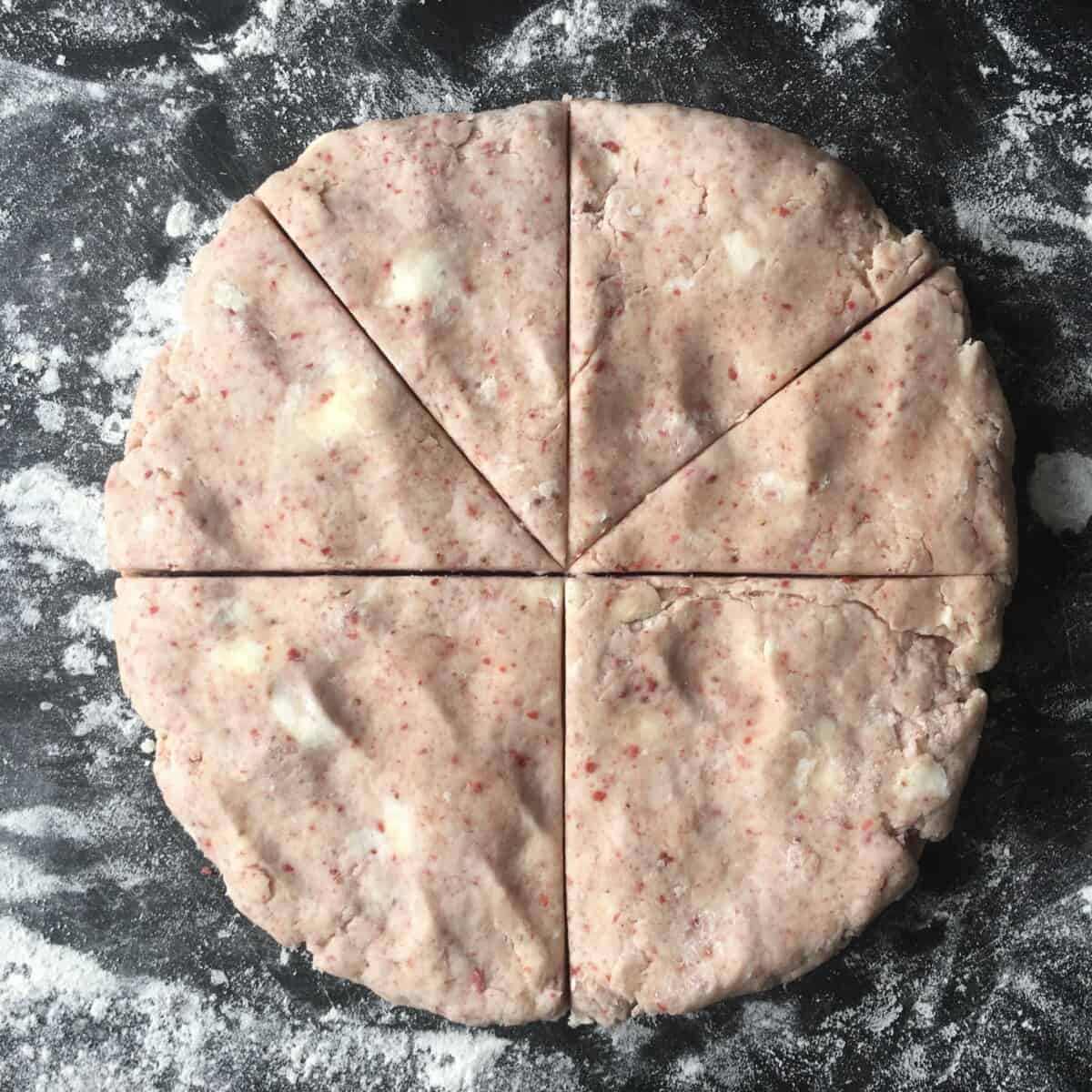 very pink strawberry scone dough with large flecks of butter throughout all in a lightly flattened round disc shape with the first triangular scone shapes cut and 2 of the quarters left without any cuts