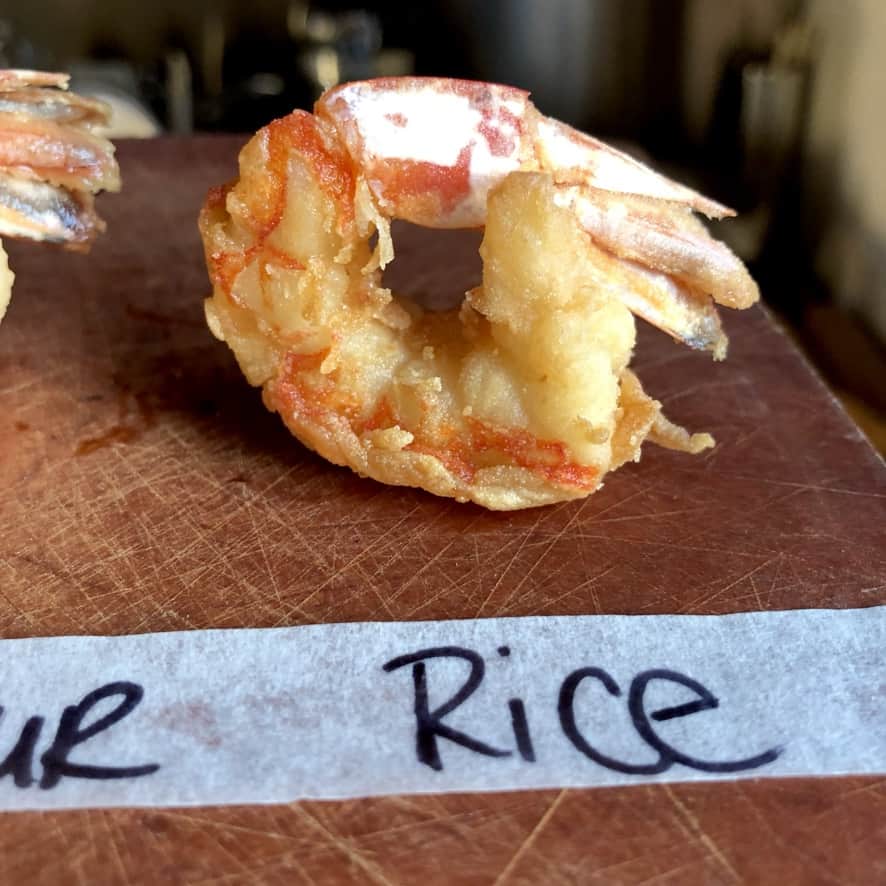 single fried shrimp with masking tape on the cutting board in front of it with black sharpied "rice flour" written on it