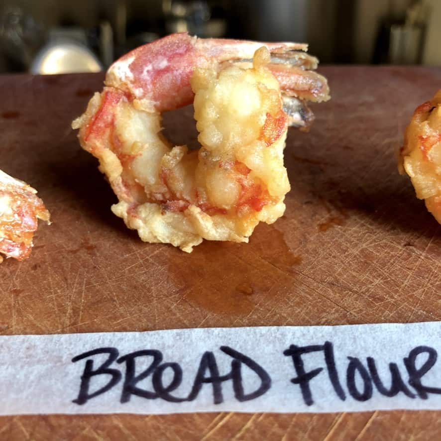 single fried shrimp with masking tape on the cutting board in front of it with black sharpied "bread flour" written on it