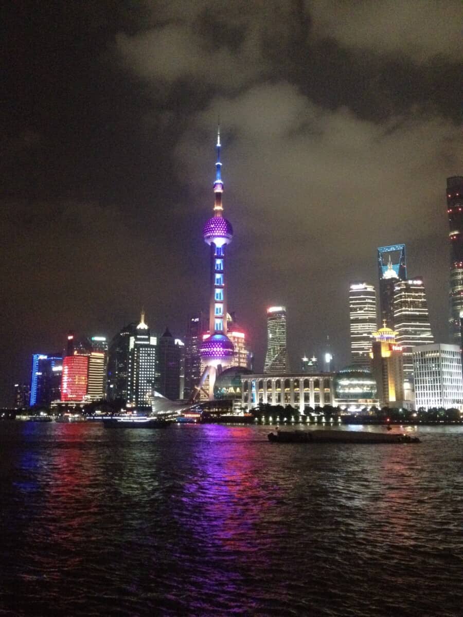 a view of the Bund taken from one side of the river in shanghai with neon lights reflecting off of the water from the iconic skyline above the Huangpu River