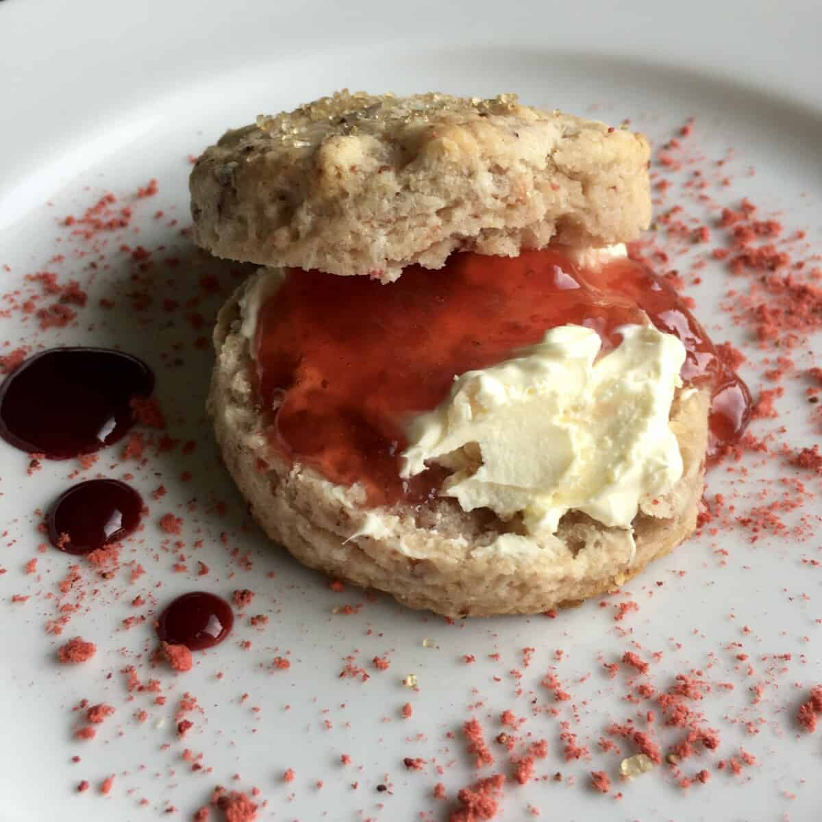 closeup a beautiful slightly pink'ish with strawberry flecks scone that's been split in half, clotted cream added, topped with homemade strawberry jam and the plate sprinkled with freeze-dried strawberry powder with strawberry coulis dots off to the side