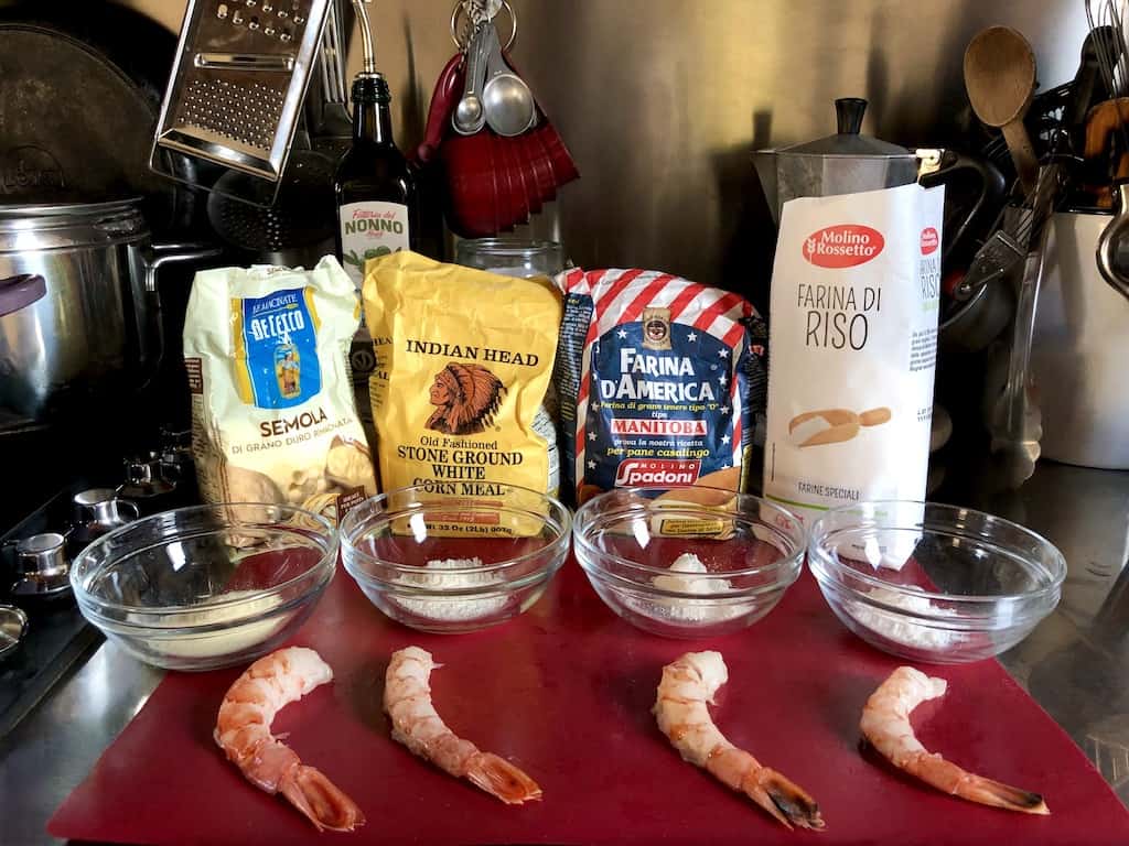 4 bags (3 bags of various types of flour + 1 bag of stoneground cornmeal) each with a small glass prep bowl in front of each bag containing a small amount of the respective flour or cornmeal and 4 shrimp lying one in front of each bowl