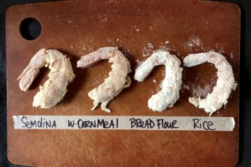 four Argentinian Red Shrimp dusted each with a different type of flour or cornmeal on an Epicurean Cutting board and a line of masking tape below the lined up shrimp indicating what has been used on each shrimp in the lineup