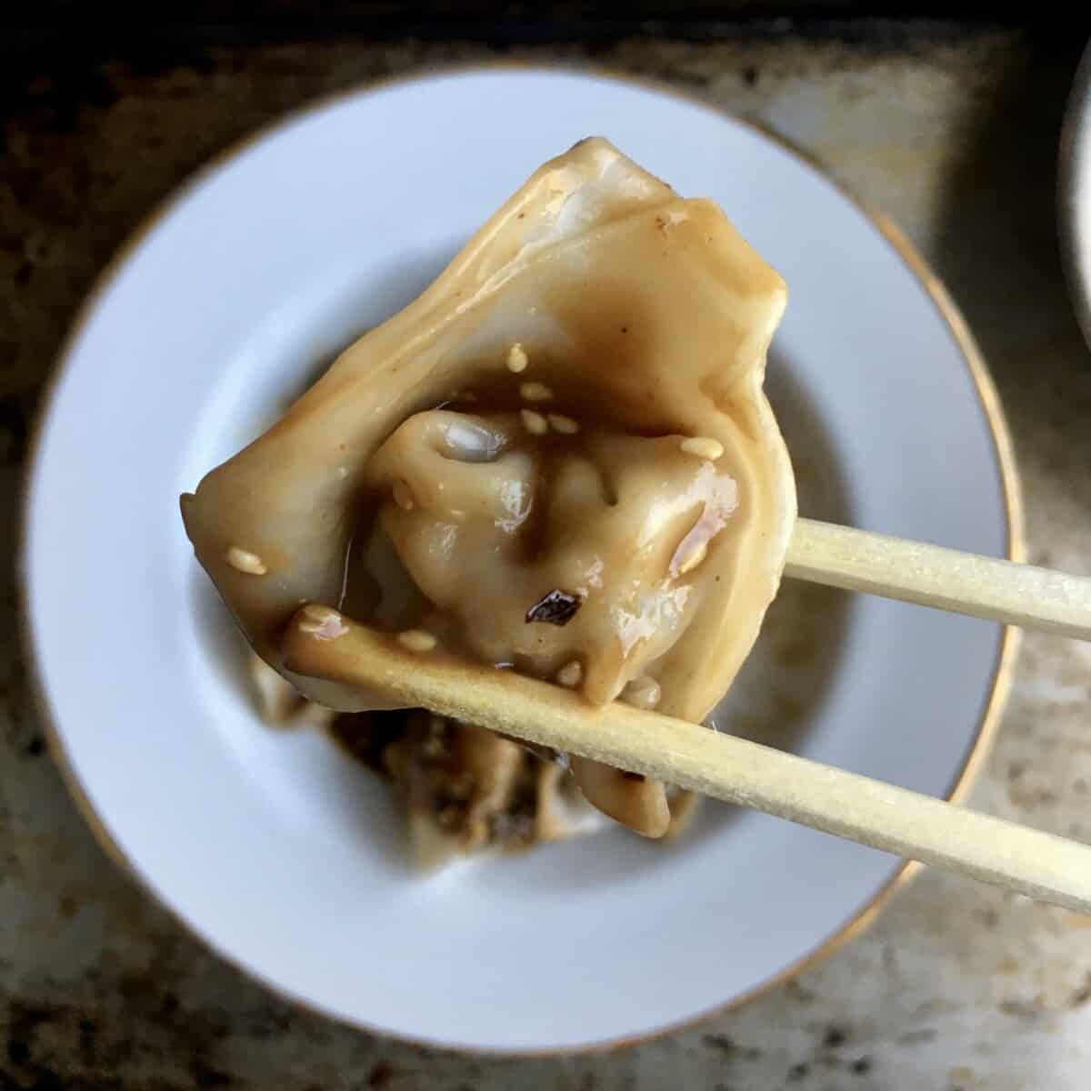 a homemade Sichuan pork wonton covered in sauce being held by a pair of chopsticks above a bowl of hot wontons