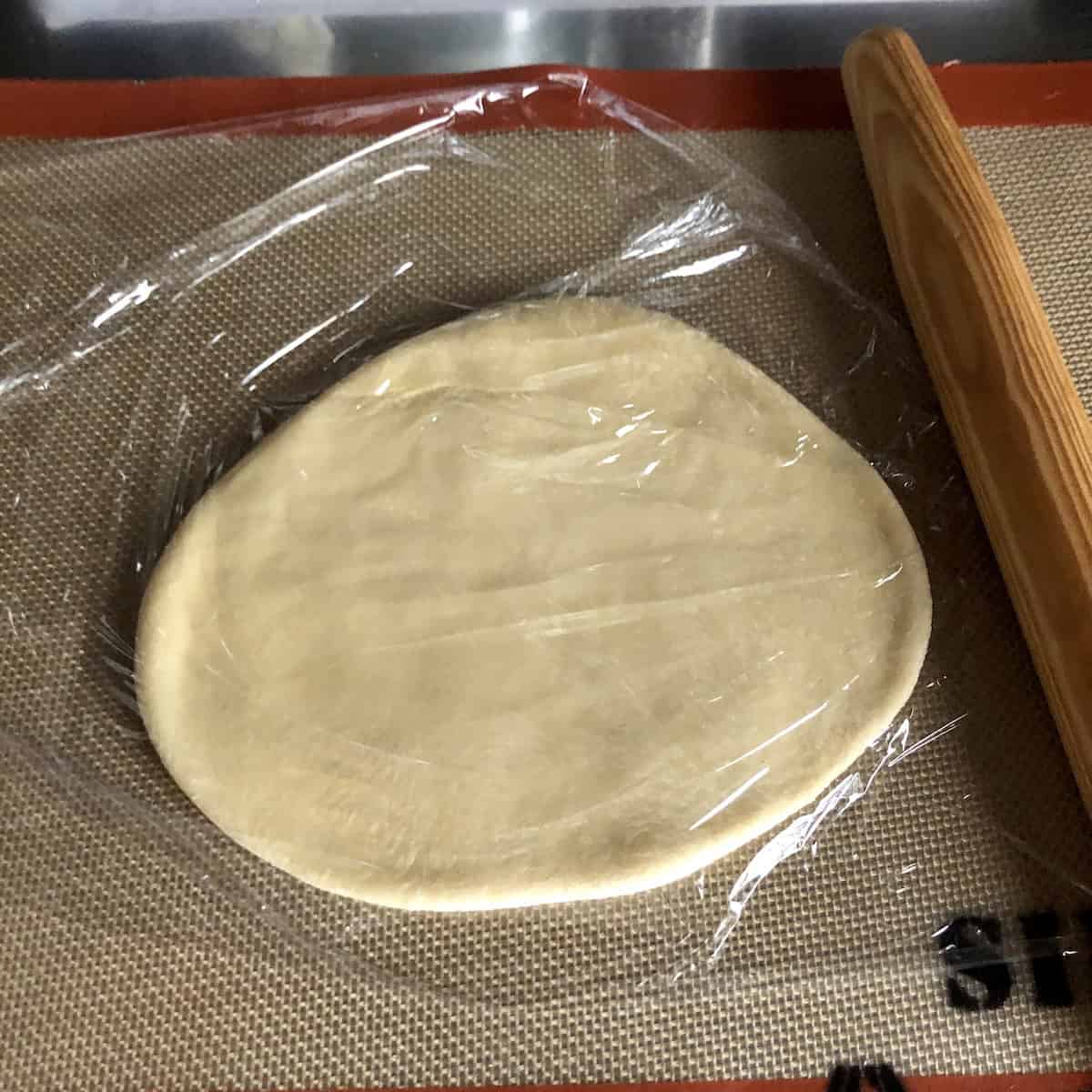 A single portion of wonton wrapper dough covered with cling film to let the gluten relax before rolling, homemade wonton wrapper recipe resting step.
