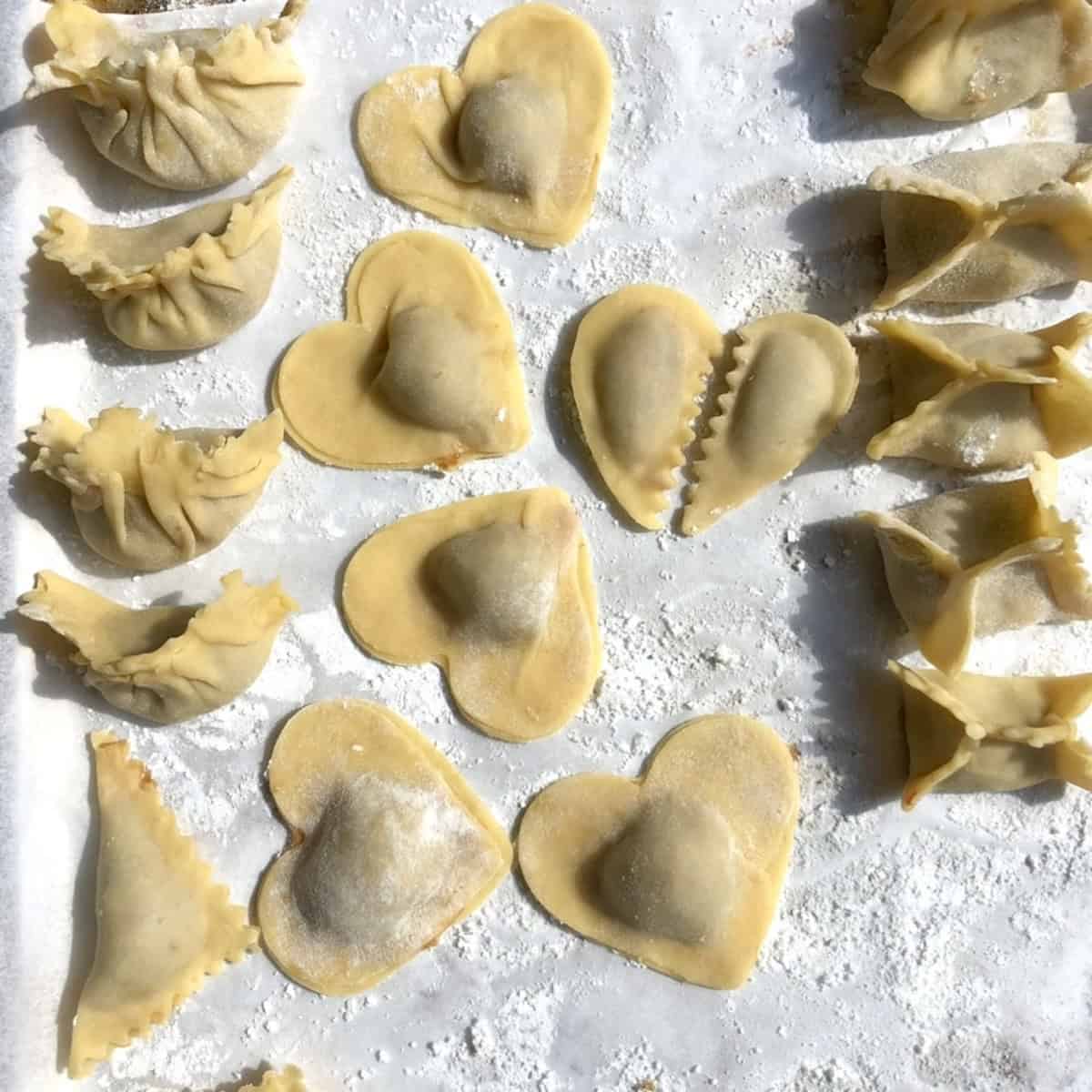 A floured tray of raw shaped wontons including heart-shaped, pleated dumpling, and traditional folded wontons made with homemade wonton wrappers ready to cook.