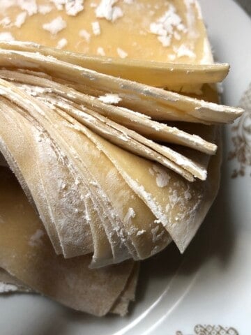 Holding a stack of homemade wonton wrappers fanned out to show the thickness and sturdiness, wonton wrapper recipe for wonton skins that hold up to filling and frying.