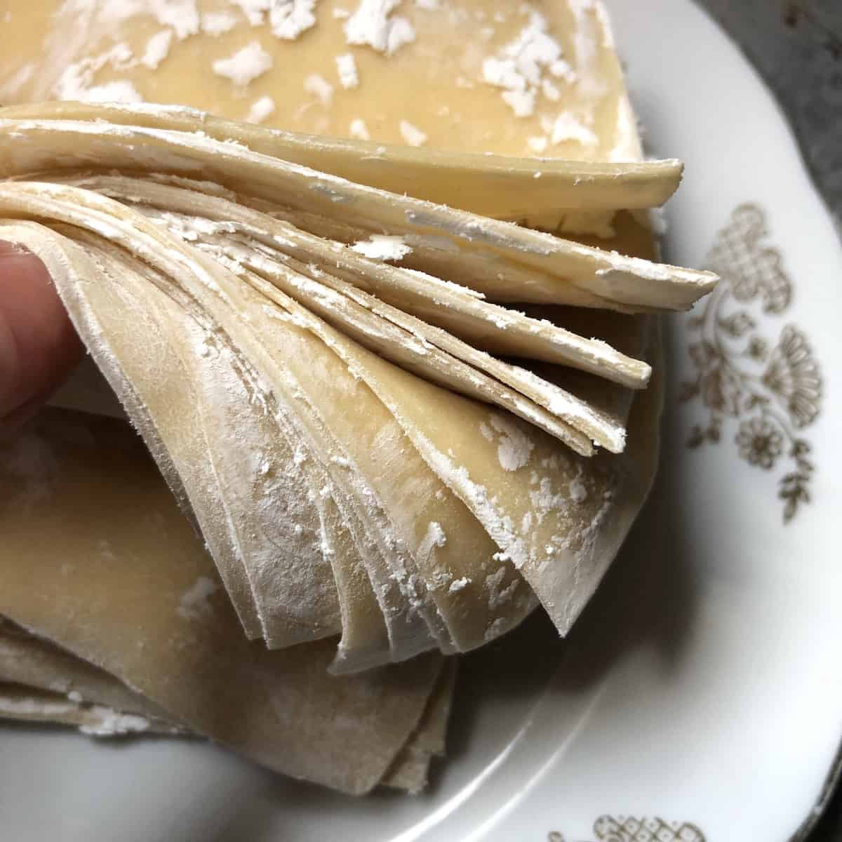Holding a stack of homemade wonton wrappers fanned out to show the thickness and sturdiness, wonton wrapper recipe for wonton skins that hold up to filling and frying.