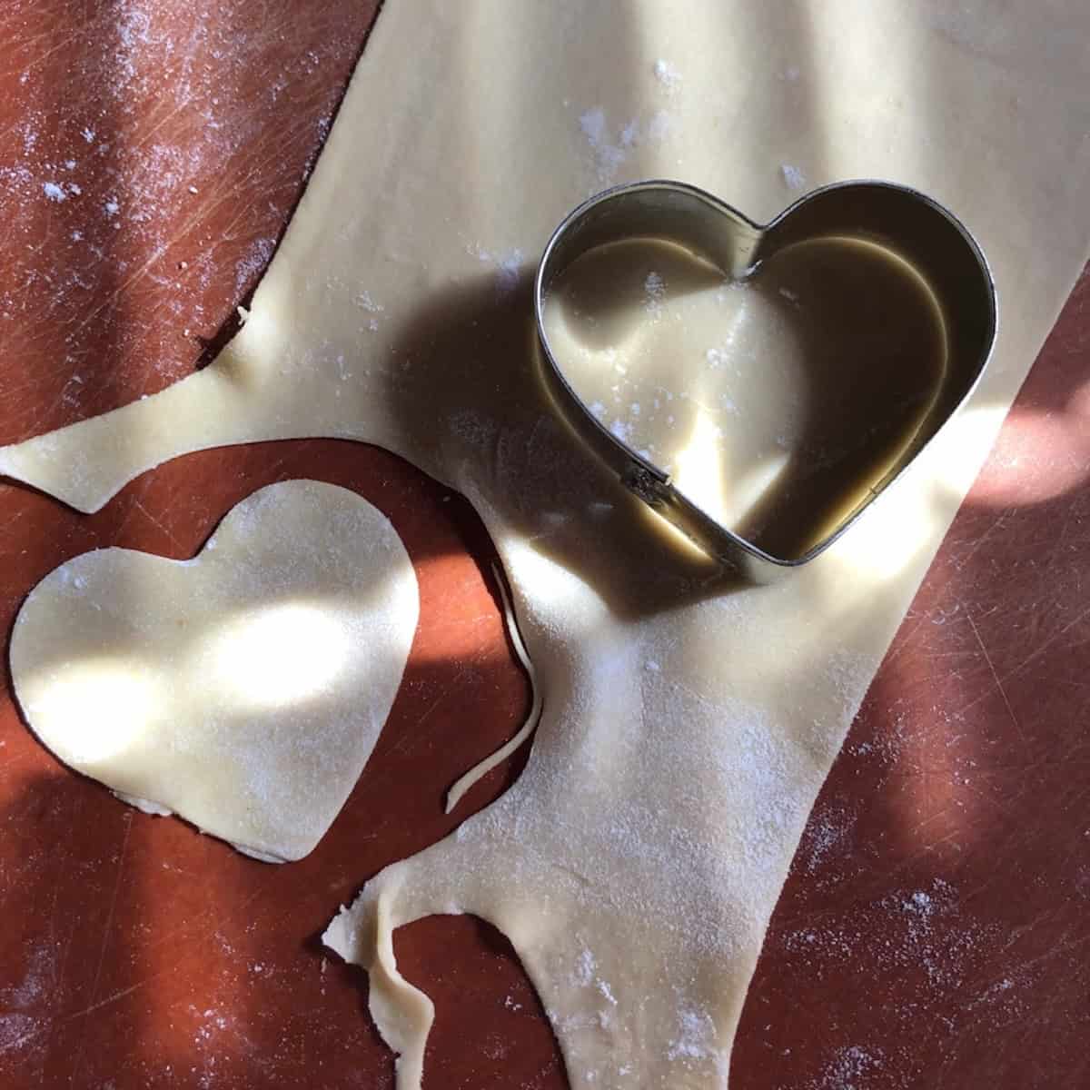 A heart-shaped cookie cutter cutting out a heart from rolled wonton wrapper dough for Valentine's Day wontons, how to make wonton wrappers in different shapes from scratch.
