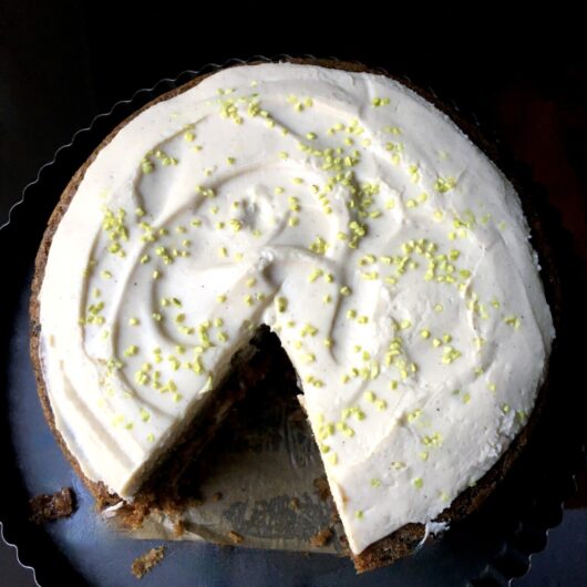 a carrot cake with cream cheese frosting and one slice removed with freeze-dried pistachios sprinkled on top