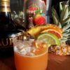 The Zio Renato in a Reidel water glass with a garnish of lime, blood orange, pineapple, and strawberry with strawberry sugar rimming the glass and coconut pieces floating on top of the orange-coral colored cocktail with the pineapple top and glass liquor bottles in the background