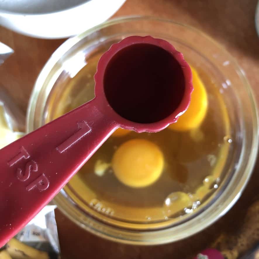 a teaspoon with vanilla extract being held above a small glass prep bowl with 3 cracked eggs in it