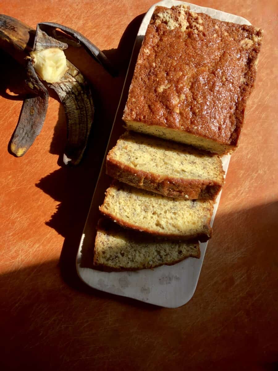 A moist banana bread loaf with 3 slices cut and a half of a banana turned upside down next to the marble tray.