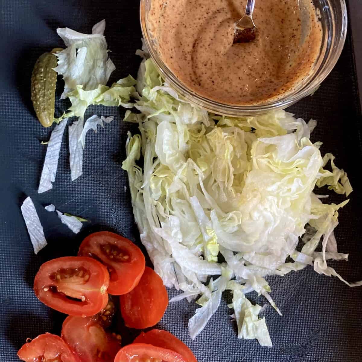 homemade smoky shack sauce with a small cornichon pickle to the left, shredded ice berg lettuce, and halved grape tomatoes
