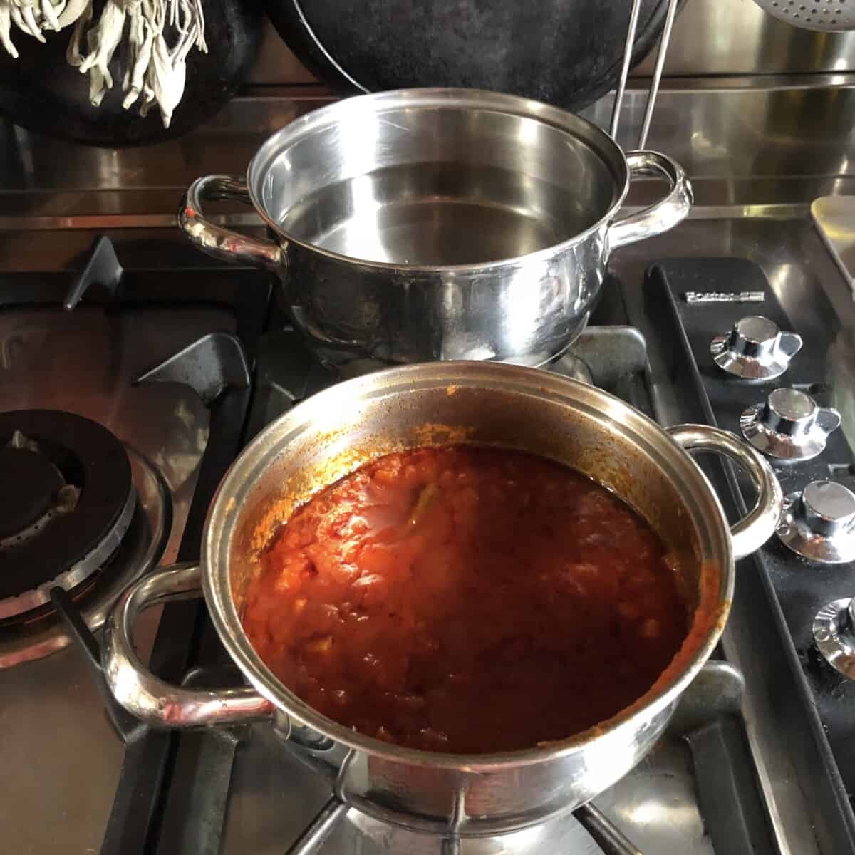 a pot of boiling water on the stove with a pot of homemade tomato pasta sauce on the burner in front