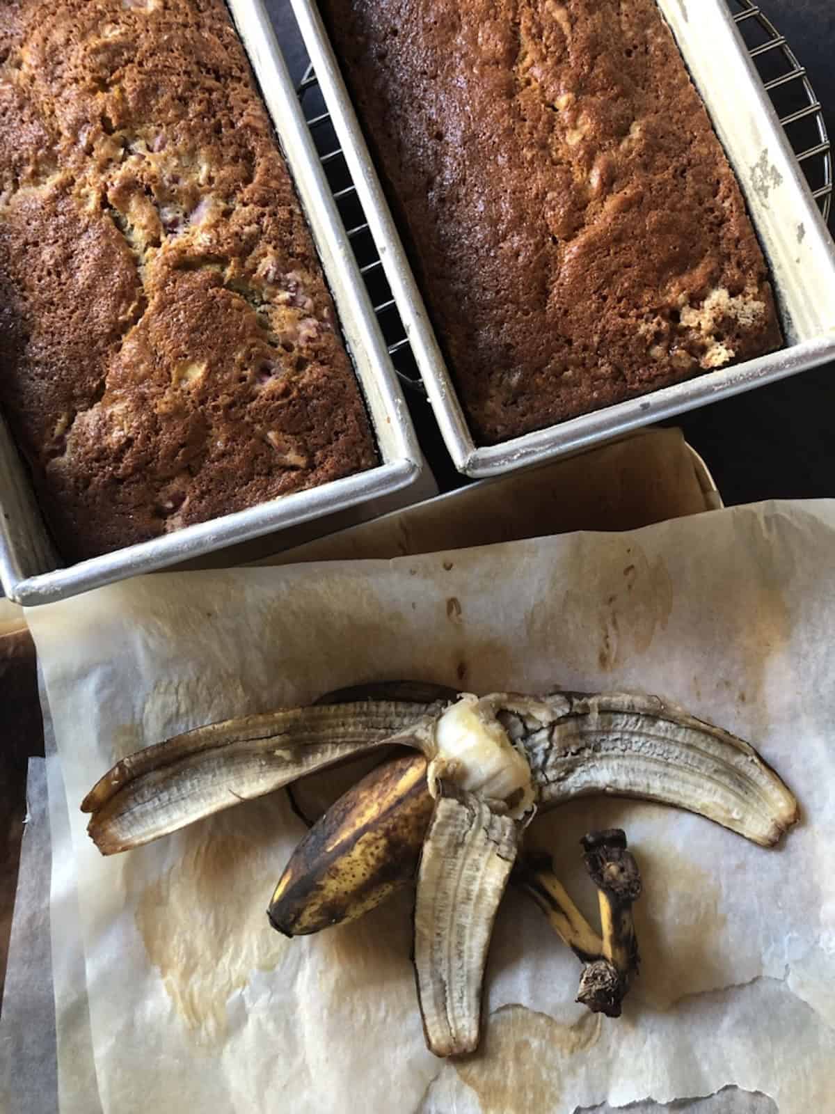 Two loaves of banana strawberry bread with fresh diced strawberries cooling in their loaf pans on a wire rack.
